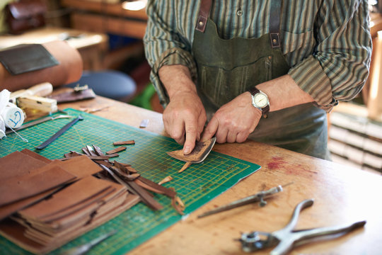 Male worker in leather workshop, checking sharpness of knife on leather, mid section