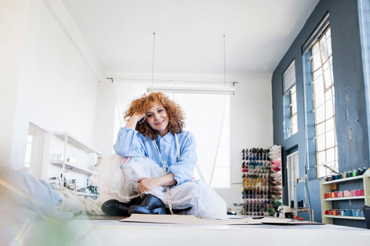 Fashion designer sitting cross-legged on desk looking at camera smiling