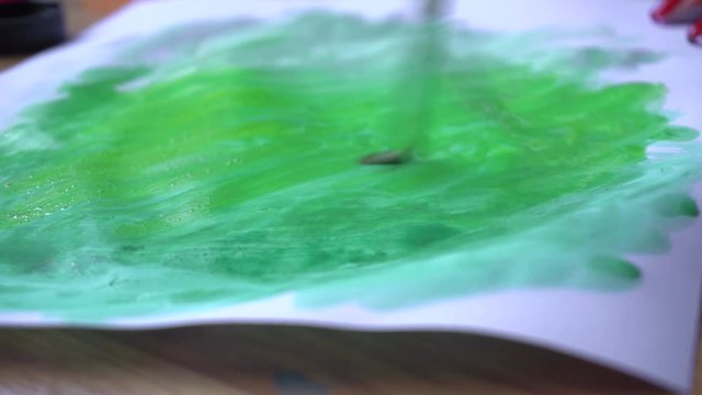a girl paints with a brush and green paint on a sheet of paper closeup