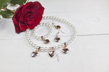 Pearl necklace, bracelet and earring with golden hearts and red rose on white wood