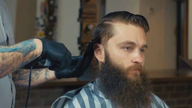 Unparalleled barber with a beard and a tattoo is cutting the hair of his client in the barbershop. He is using a cutting comb and a hair clipper.