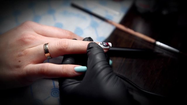 Manicurist in black gloves gets a top coating on the nail with drawing of a penguin shot close-up under bright light.