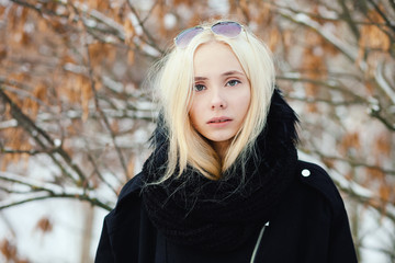 Close up winter portrait: young blonde woman dressed in a warm woolen jacket posing outside in a snowy city park with foliage background