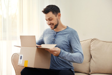 Handsome young man opening box with parcel while sitting on sofa at home