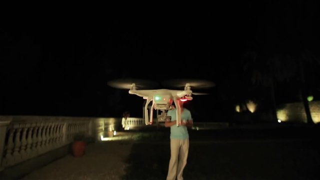 Drone quadcopter taking off from ground to night sky with operator controlling it side view. Man operates drone lighting up using remote controller. Guy pilots rc quadrocopter at black sky background