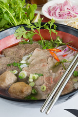 A bowl of traditional vietnamese beef and chicken pho street food, surrounded by fresh herbs. Pho is a vietnamese noodle soup consisting of meat, herbs, chilli, coriander and dumplings.