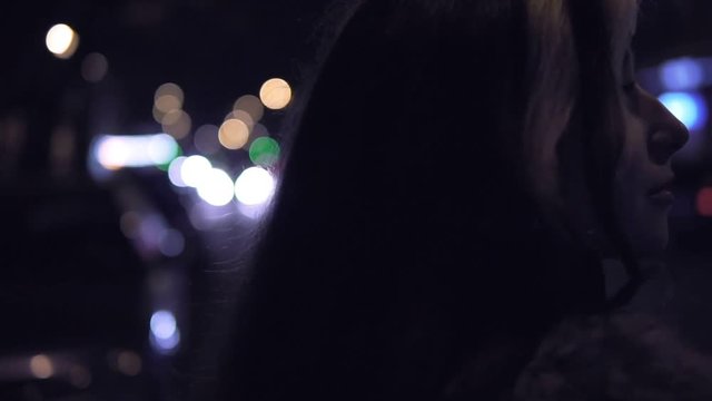 Young brunette woman stands on the street at night. City traffic at the background