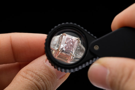 a brilliant pink diamond ring is being held by hand and looked through a loupe