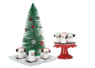 Marshmallow s'more men with ear muffs sitting on a chocolate bar square. Festive Christmas holiday snacks served on pedestal next to a miniature tree