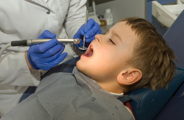 Dentist is treating  teeth of five y.o. boy sitting in the dentist chair under the medical lamp light with wide opened mouth and closed eyes - 131270139