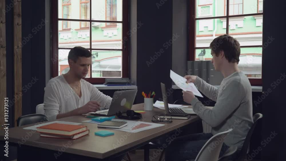 Aufkleber Young professional start up team working in modern office. Caucasian men computing on laptop and discussing project. Handsome man wearing in casual clothes talking with friend at work. Smiling workers