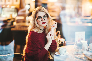 Beautiful young blond woman with glasses sitting in a cafe and drink coffee, it can be seen through the cafe window