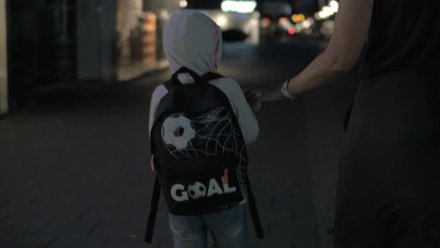 Slow motion steadicam shot of boy with backpack and his mom walking in night street. Mother teasing child with a toy