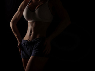Fototapeta premium Young athletic and sexy woman in a white sports shirt and blue shorts in the studio on a black background shows strong biceps and a flat belly with the perfect abdominal muscles, dark black background