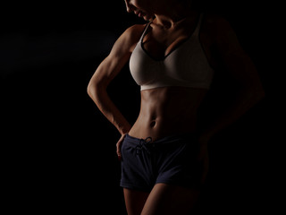 Obraz na płótnie Canvas Young athletic and sexy woman in a white sports shirt and blue shorts in the studio on a black background shows strong biceps and a flat belly with the perfect abdominal muscles, dark black background