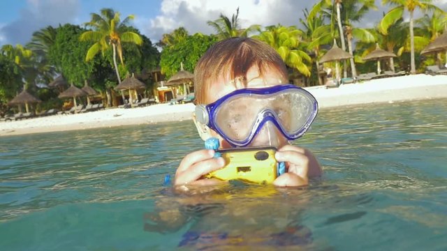 Little boy in snorkel mask taking pictures with camera in waterproof case over and under the water. Vacation on the coast in tropics