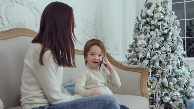Little boy talking on the phone describing presents he got for Christmas sitting on the sofa with his mom