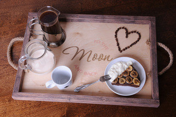 A piece of chocolate cake with banana, coffee in french press and pitcher with cream on a wooden...