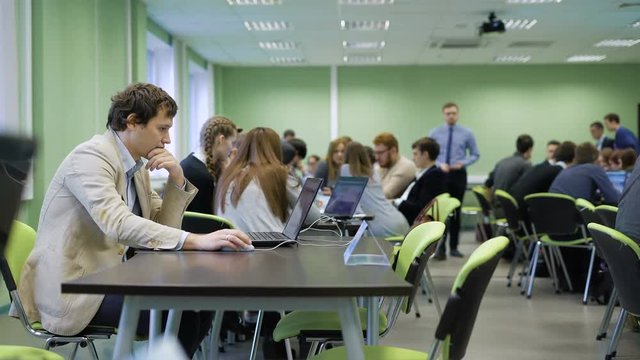 Organizer of business game sitting at the desk in the classroom and using laptop during students at the room are doing practice and playing their roles ccording to the rules of the game.
