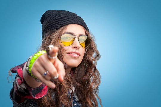 Closeup portrait of beautiful trendy hipster girl with curly hair pointing finger at you and smiling wearing red checkered shirt,denim vest and black beanie hat on blue background.Youth style,fashion.