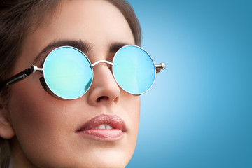 Close-up face portrait of young beautiful woman with perfect skin in round sunglasses looking up on blue background. Beauty face make-up.