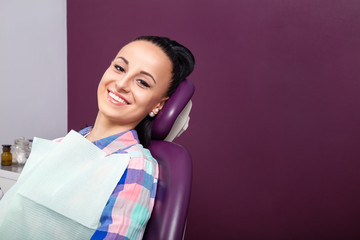 Young woman patient in checkered shirt with perfect straight white teeth waiting for dentist in dental chair and smiling relaxed, ready for a check-up. Beautiful woman smile