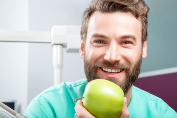 Portrait of young handsome male doctor with beard smiling with perfect straight white teeth holding green fresh ripe apple. Face expressions, emotion, healthcare, medicine.