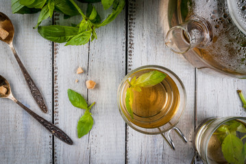 Basil herbal tea in a transparent cup and teapot, spoon with cane sugar, a bunch of fresh basil. On a white wooden table. Copy space 