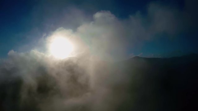 Aerial bird-eye view flight through clouds flying towards bright sun and blue sky background also showing mountain landscape below beautiful scene drone stable forward bird view flight relaxing 4k