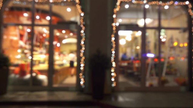 Blurred windows of coffee house with glowing lights. People are walking the street near cafe at night.