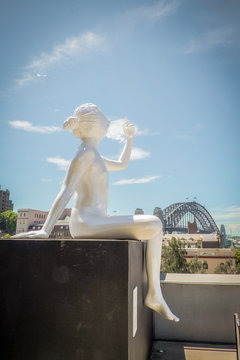 the pure white color of sculpture shown as the lady sitting and point to the blue sky with the relaxing act and the harbour bridge is located far away from the sculpture 