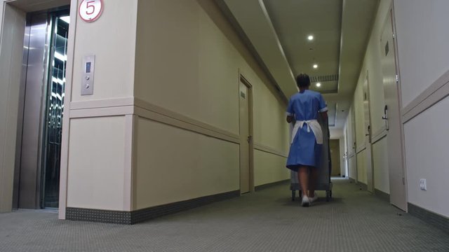 Low angle shot of female Latin-American housekeeper in uniform getting out of elevator and pushing trolley along hall in hotel