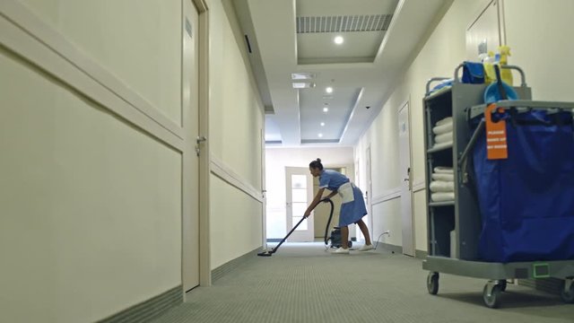 PAN of Latin-American cleaning service lady in uniform vacuuming carpet in hotel hall 