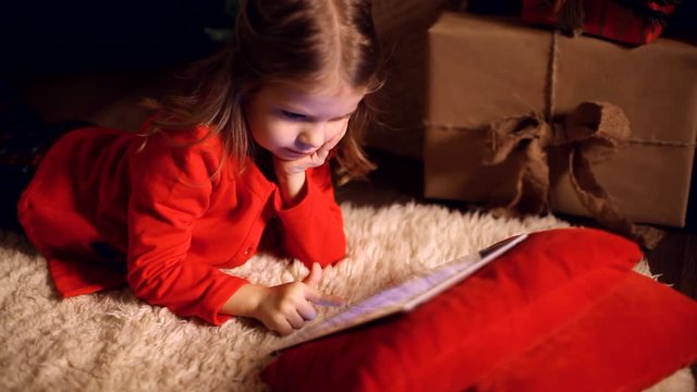 Little girl lying in carpet with presents around using tablet on red pillow.