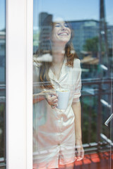 Young woman standing behind glass of balcony door, drinking morning coffee