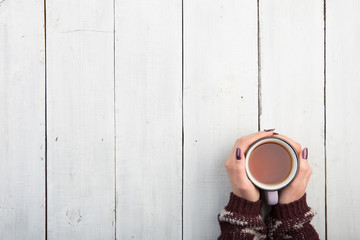 Woman's hands in sweater holding cup of tea on the white wooden table