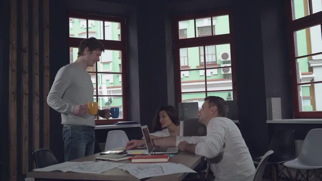 tired employees fall asleep at work. Young businessman and business woman sleeping at the working place. Happy smiling man associate entering into the room and bring coffee. Handsome workers holding
