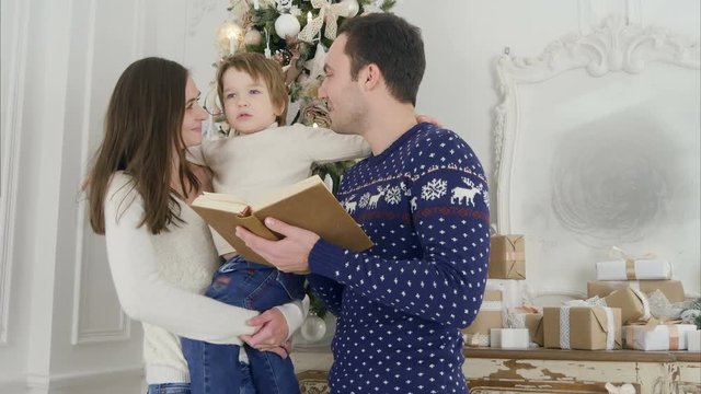 Happy father reading a Xmas tale while mother holding their cheerful son in front of the Christmas tree
