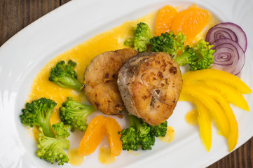 Delicious fillets fish roasted in the frying pan. Posted with tangerine sauce, pepper and slices of boiled broccoli al dente. Wooden background. Top view. Close-up
