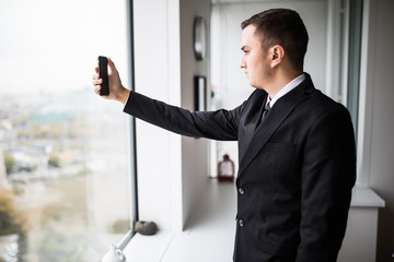 handsome young businessman in a stylish modern office space with large windows, taking selfie in phone