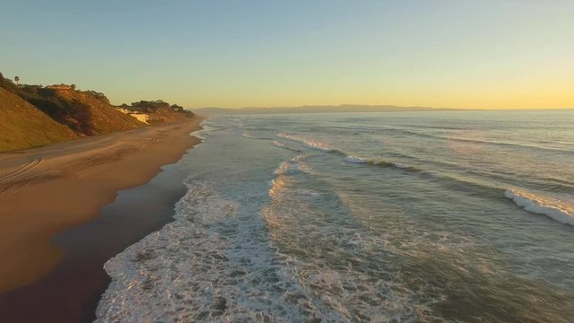 Aerial flying shot down a beach in California over the sand and water. Beach houses in the distance. Sunset time with clear blue skies and golden light. 