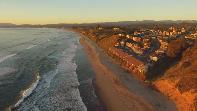 Slow aerial shot moving forward over the beach in California overlooking beach houses during sunset. Clear blue skies, blue water, and golden light. 