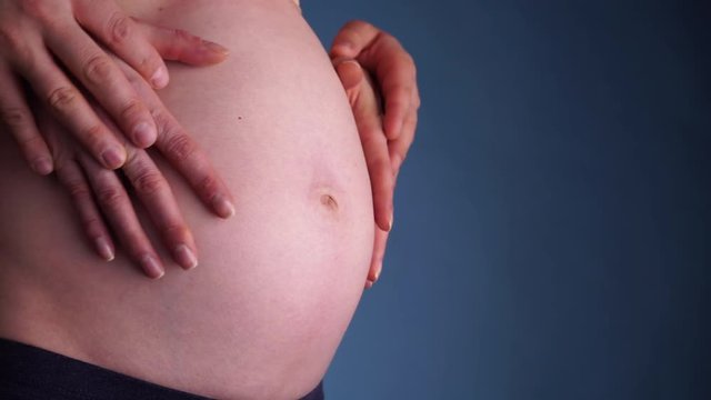 Couple making heart shape on the pregnant naked belly with their hands, closeup on dark navy blue. Concept of pregnancy, expecting a baby, love, parenthood. 4K ProRes HQ codec