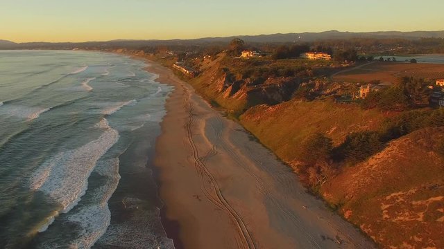 Aerial flying shot at the beach in California over sand and water, with beach houses in the distance. Golden light of sunset. Clear skies, vehicle tracks in the sand. 
