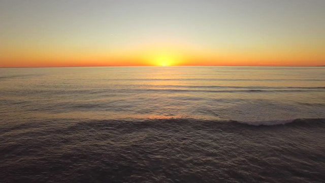 Aerial shot flying backwards over the ocean in California during sunset. Sharp horizon line with golden light. Rises at the to end reveal beach and waves. Clear skies, vibrant colors. 