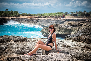 Portrait of young and sexy woman on the rocks near the wild ocean. Storm, huge waves coming and splashing. Tropical island Nusa Lembongan, Indonesia, Asia. The name of the place is Devils Tear.