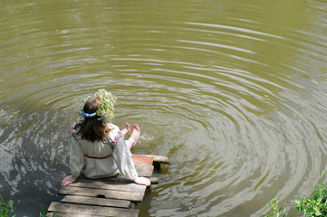 Молодая русская девушка в льняном национальном платье купает ноги в озере, сидя на деревянном мостике 