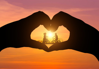        silhouette little boy and little girl  having fun riding bike on sunset  
in the middle silhouette hands heart shape 

