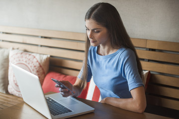 beautiful young woman is receiving a text message in her mobile cellphone device using a laptop pc while in a cafe restaurant