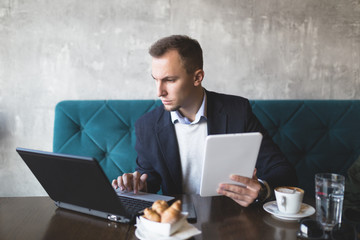 Young casually dressed business man sitting in modern office or cafe restaurant and doing something on his tablet and notebook.